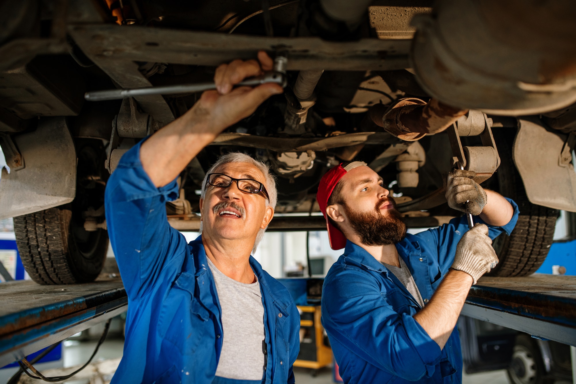 Two mechanics work underneath a vehicle, using tools to perform repairs. They are focused on their tasks, wearing blue overalls and gloves.
